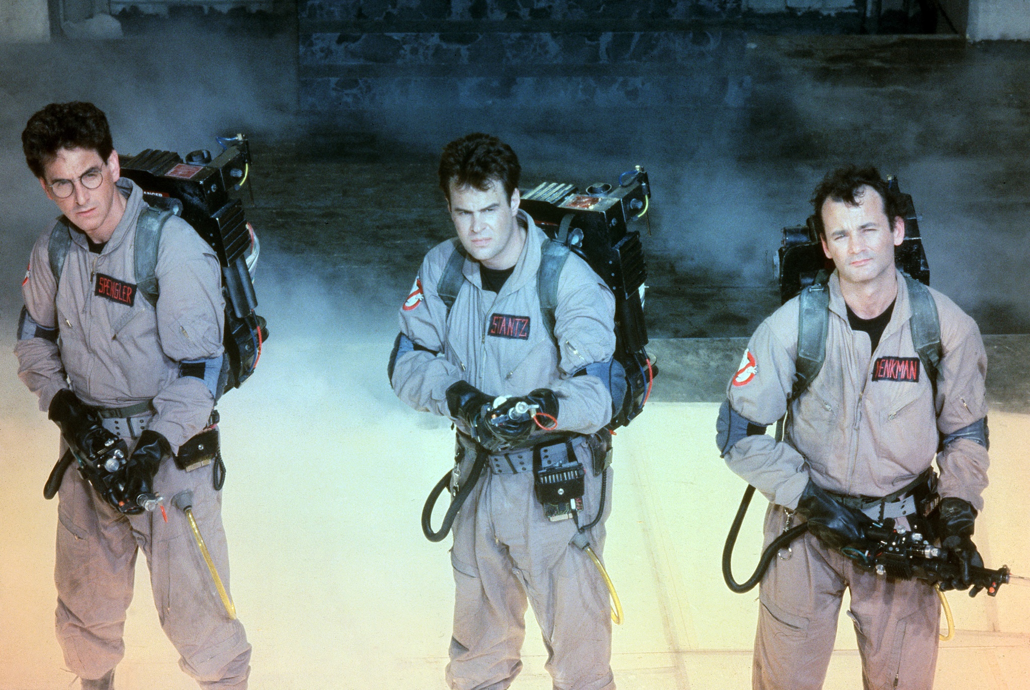 Photo of Ghostbusters. From left to right: Harold Ramis (Left); Dan Aykroyd (middle); Bill Murray (right). All are dressed in Ghostbusters one-pieces and holding ghost vacuums from film. 